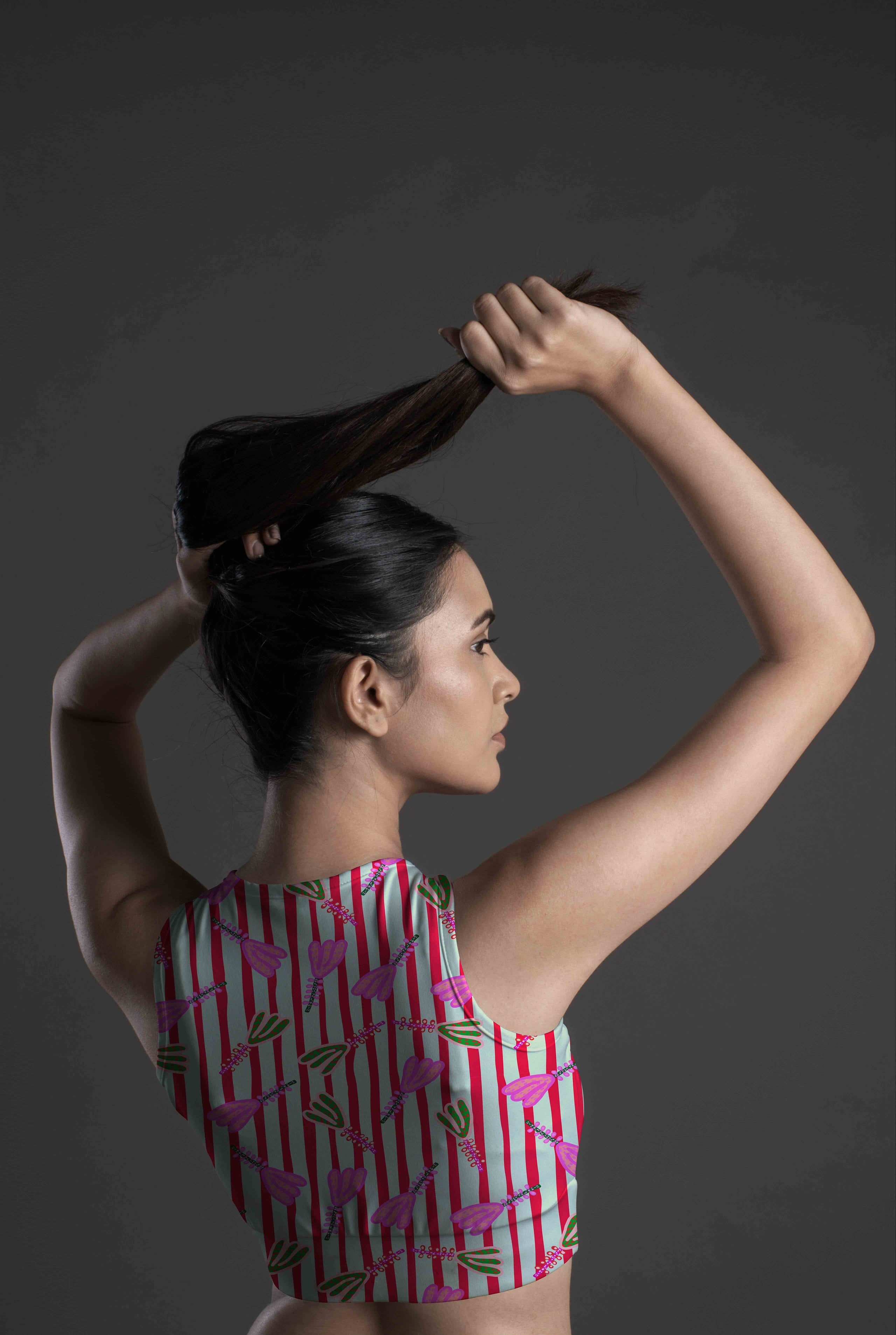A woman with long dark hair, viewed from the side and back against a dark gray background, lifts her hair with both hands while wearing the Linear Petal Sports Bra.