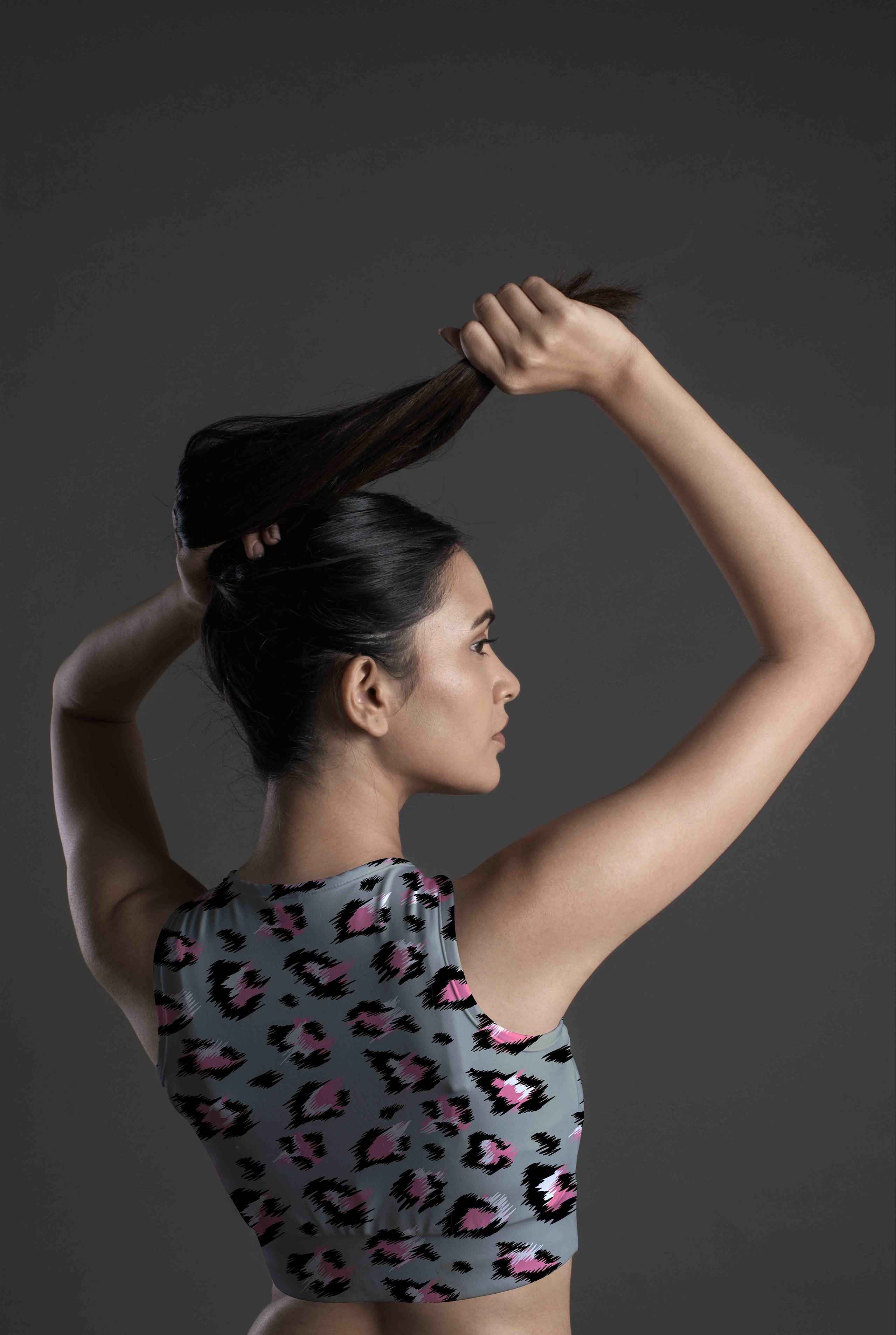 A woman with long dark hair, seen from behind in profile, lifts her hair while wearing the Wild Blush Sports Bra—a sleeveless leopard-print top featuring pink, black, and grey—against a dark plain background.