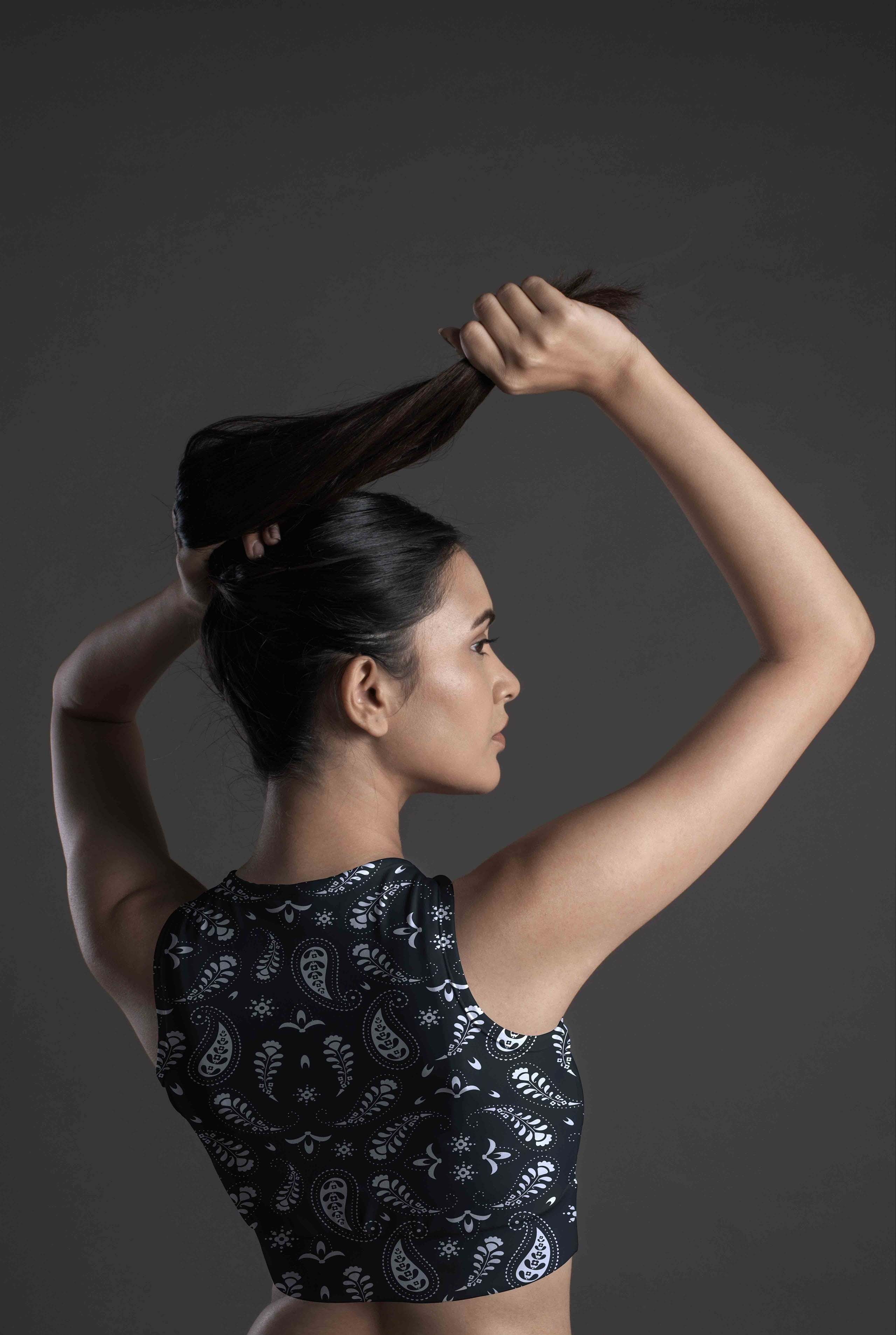 A woman with long dark hair in an Onyx Paisley Sports Bra stands sideways against a dark background, lifting her hair with both hands.
