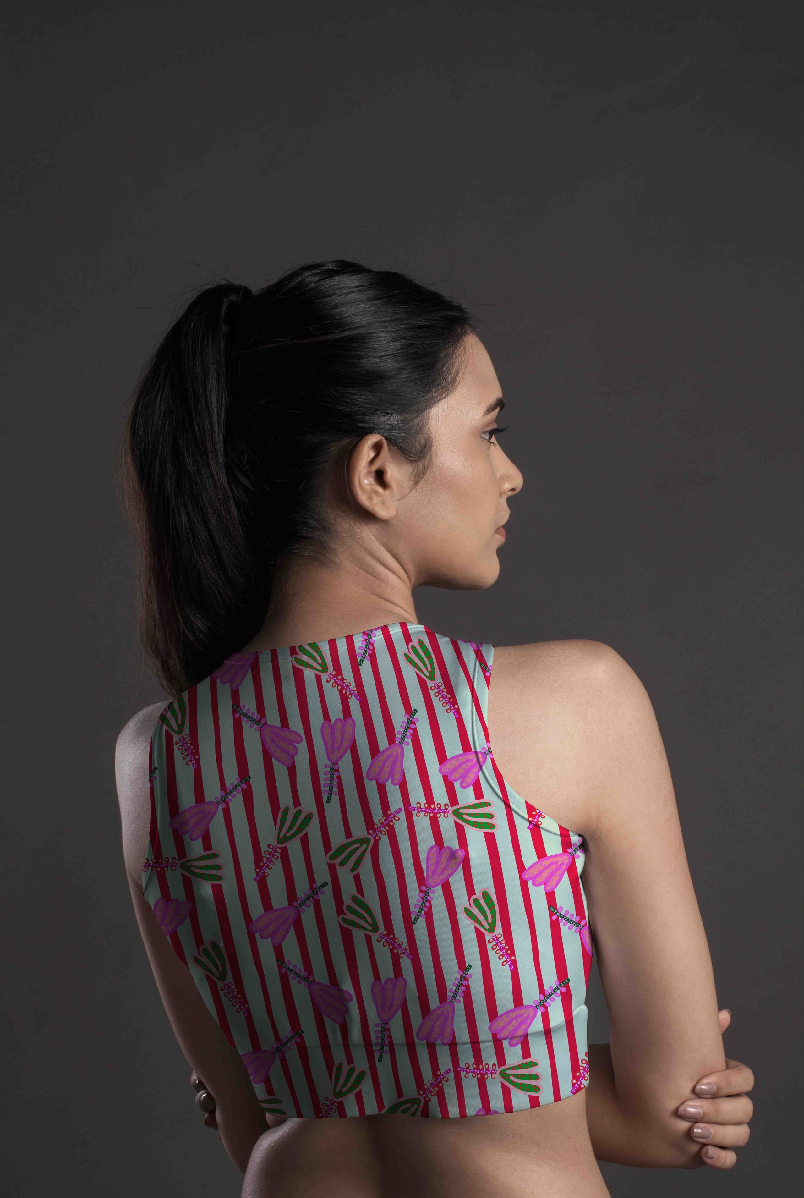 A woman with long dark hair in a ponytail, seen from the back, wears the Linear Petal Sports Bra in a pink, green, and white design on a dark background. Crafted from moisture-wicking fabric for style and comfort.