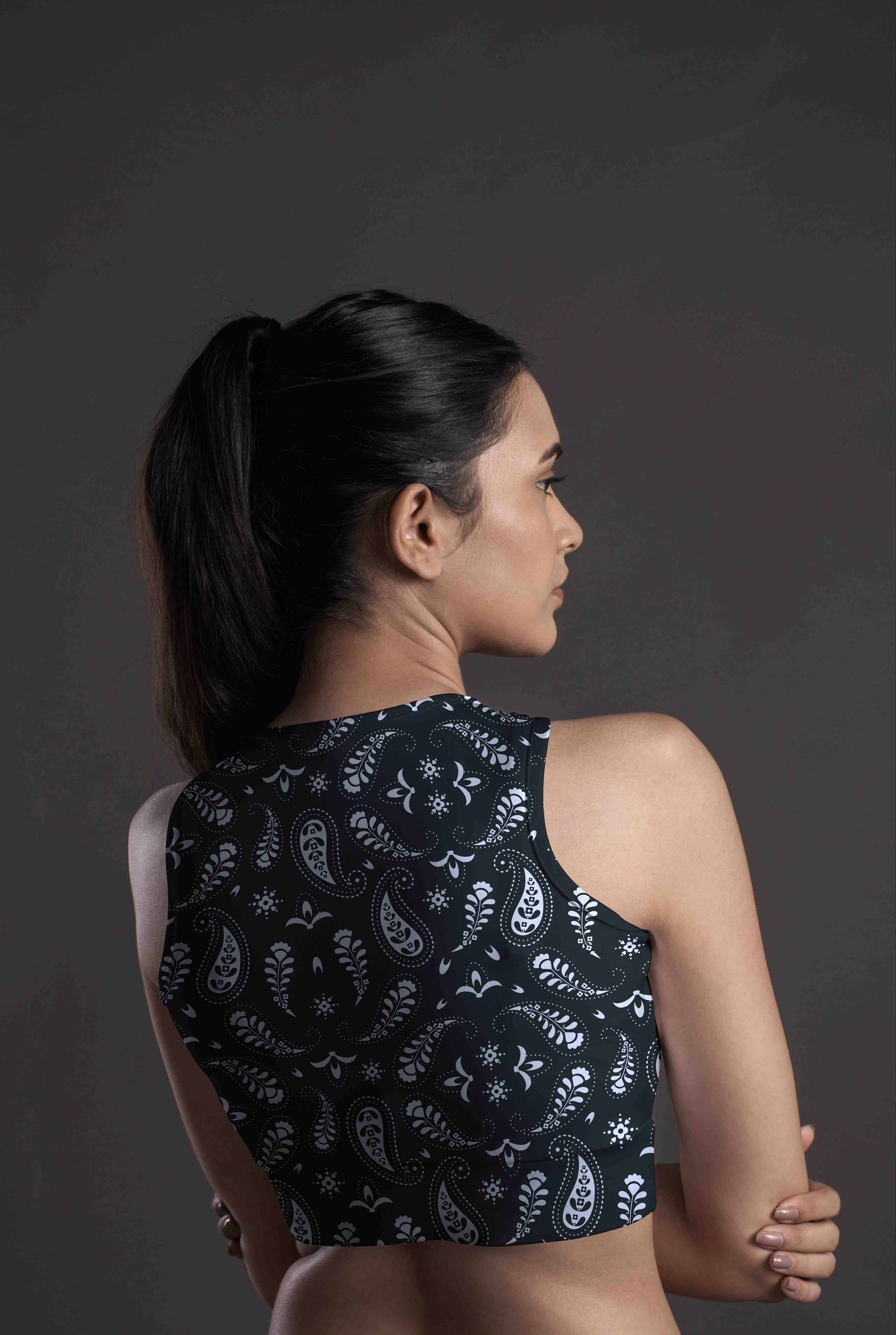 A woman with long dark hair in a ponytail, seen from behind and the side, wears the Onyx Paisley Sports Bra featuring a white paisley pattern on a dark background.