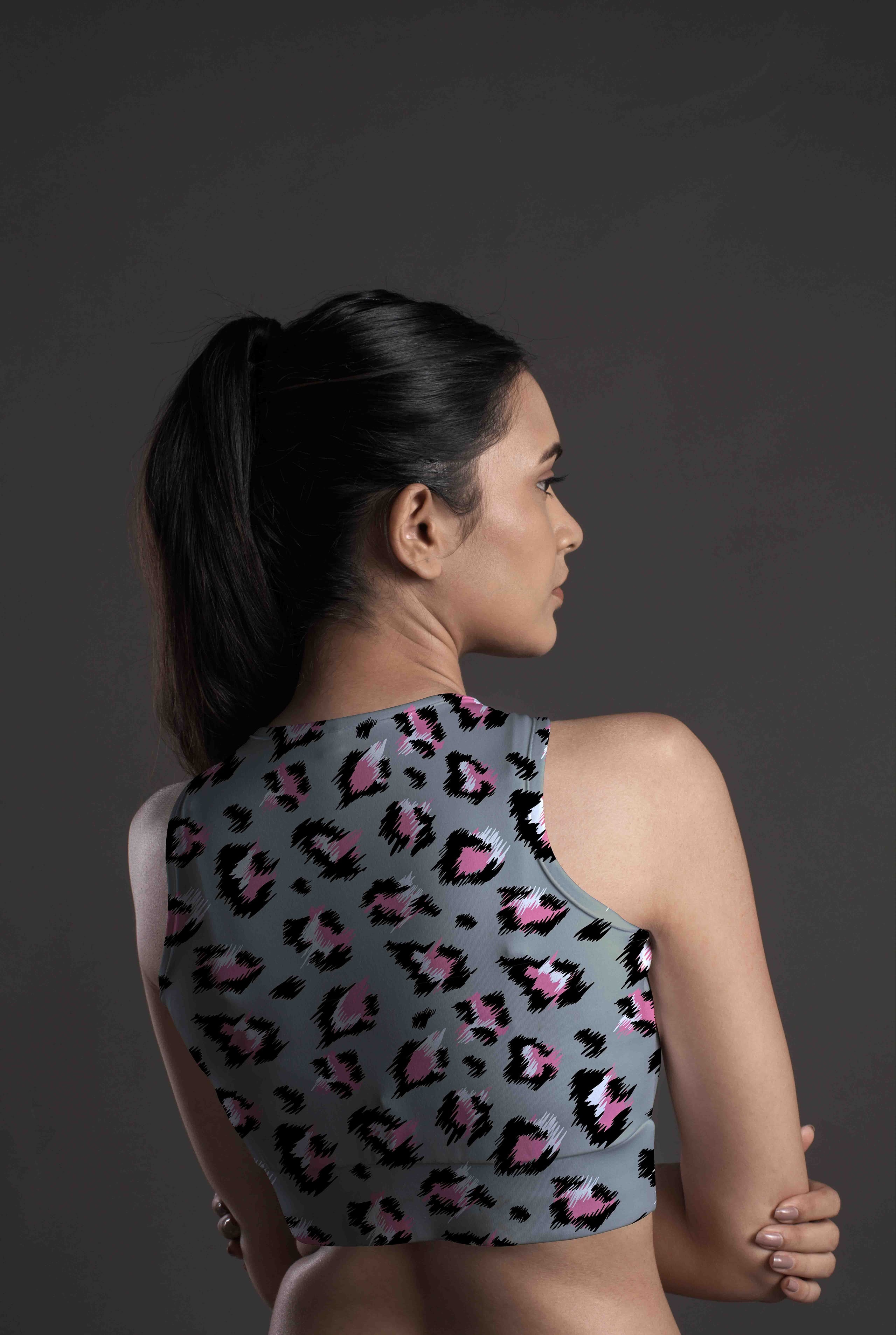 A woman with dark hair in a ponytail is seen from behind, wearing the Wild Blush Sports Bra—featuring a pink and black leopard print on moisture-wicking fabric—standing against a dark background.
